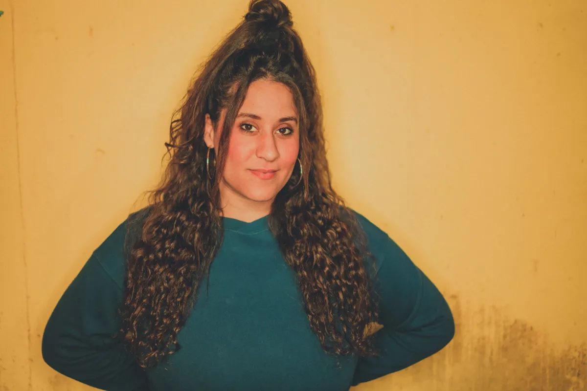 Jess Rae Ayre stands against a yellow wall and is wearing a teal sweater and a slight smile. 