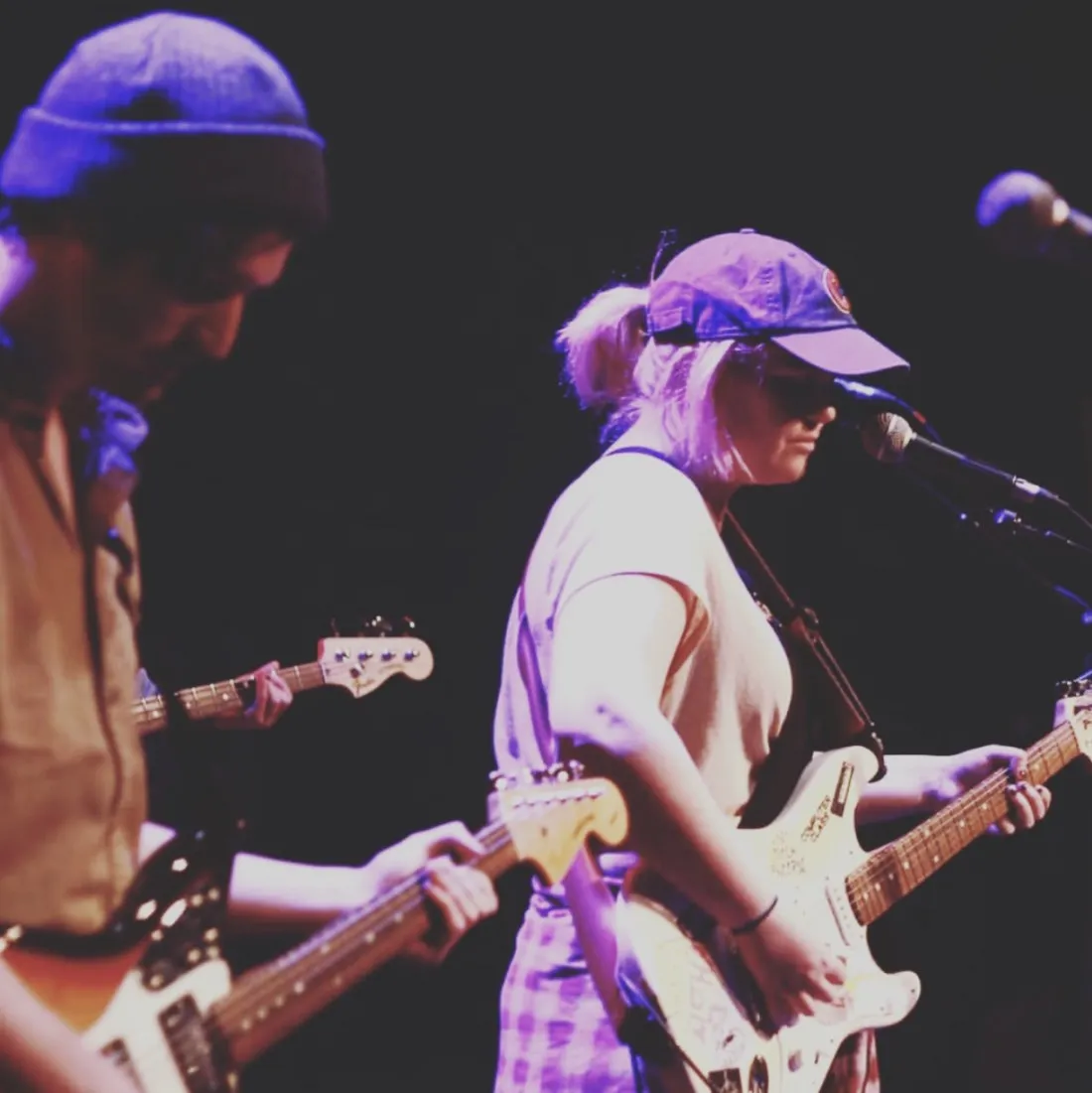 Two guitarists, one wearing a toque and the other wearing a baseball cap, play on stage under purple light. A microphone and another guitar are visible on the edges.