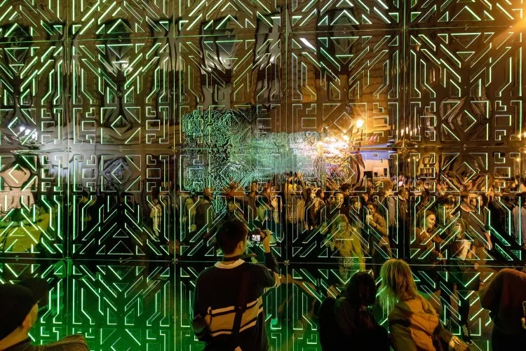 Five people stand in front of intricately patterned joined mirrors that reflect green lights and a throng of onlookers.