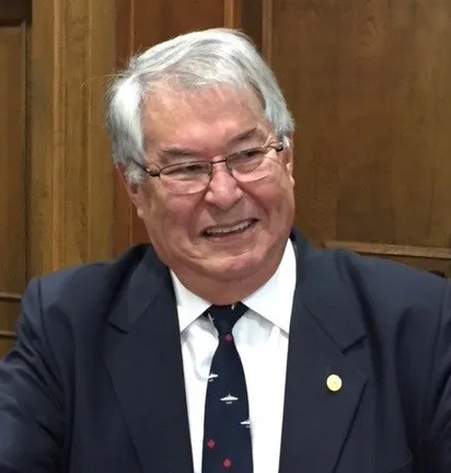 Bill Shead is an older man with white hair and glasses. He is smiling, looking to the side and wearing a dark blue suit and blue tie with white fighter jets on it.