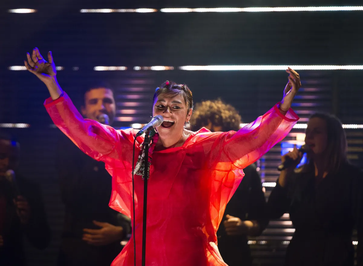 A woman singing at a microphone, gesturing widely with her arms. She wears a dark red, sheer dress with long arms.