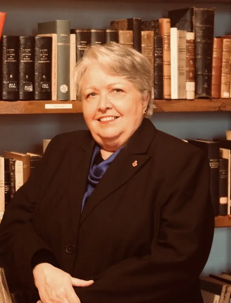 Randi Gage is an older woman with short, fair hair wearing a black blazer. She sits in front of a bookcase.