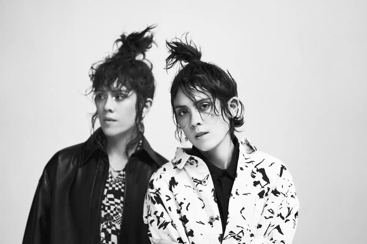 Two young people with their hair in messy topknots. One wears a black leather jacket, the other a white jacket with a pattern of black splotches.