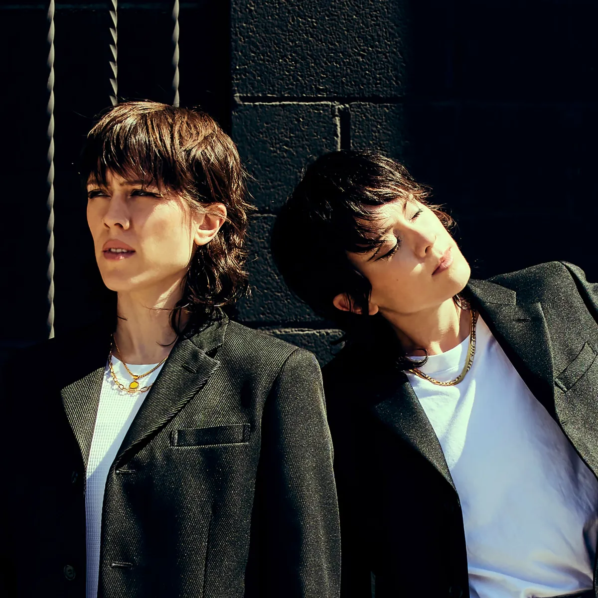 Two people wearing white shirts and black blazers pose against a black-painted cinderblock wall.