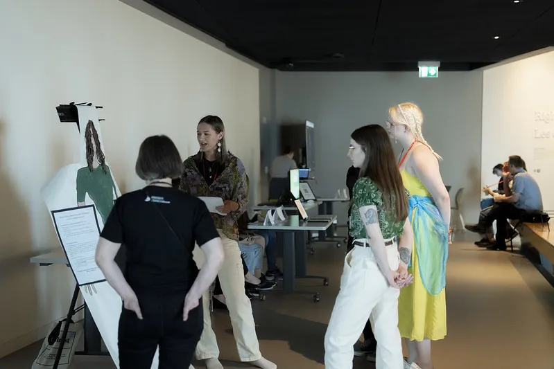 In a museum gallery, a student talks to three people next to a life-size drawing of themself.