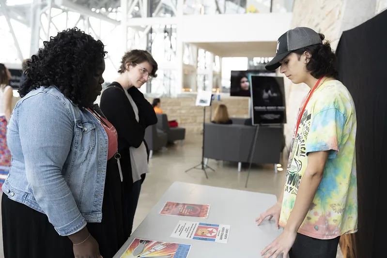 One student and two adults stand on opposite sides of a table with posters on it.