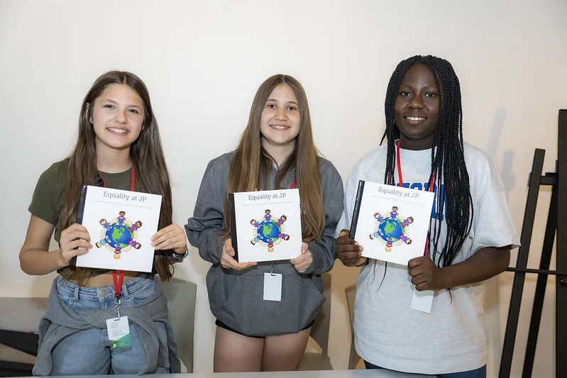Three students hold copies of a book that they've created. The book cover has a title that says "Equality at JP" and has a drawing of the earth with students all around it.