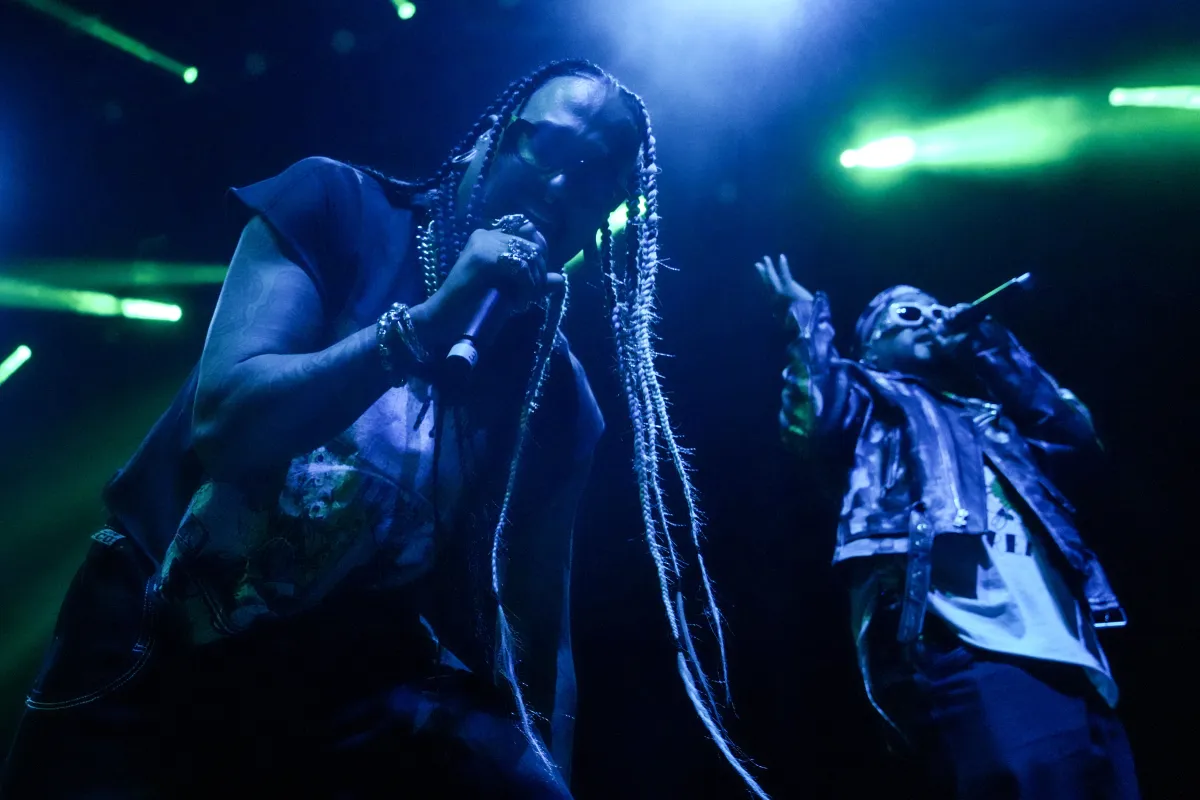 Two people holding microphones performing on stage. One is looking straight at the camera. They are bathed in blue light and with green stage lights in the background.