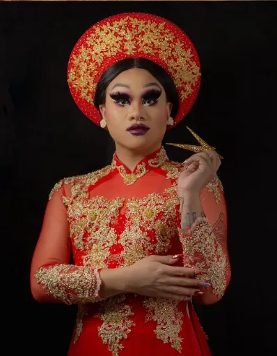 A drag artist is wearing Vietnamese cultural clothing with a matching red and gold headdress and long dress.