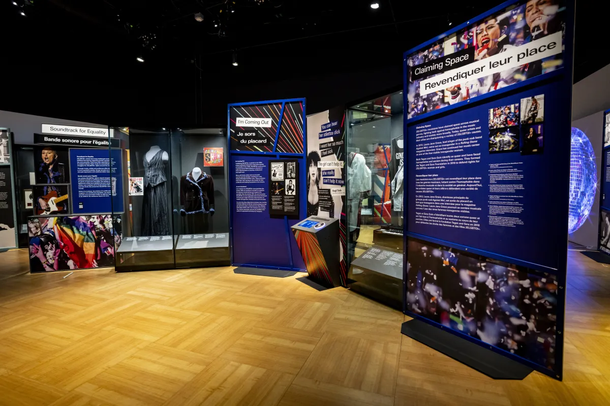 Clothing in two display cases, text and bright graphics are displayed on a standalone, curved wall with messages of 2SLGBTQI+ equality. A screen on a podium-type stand is in the centre.