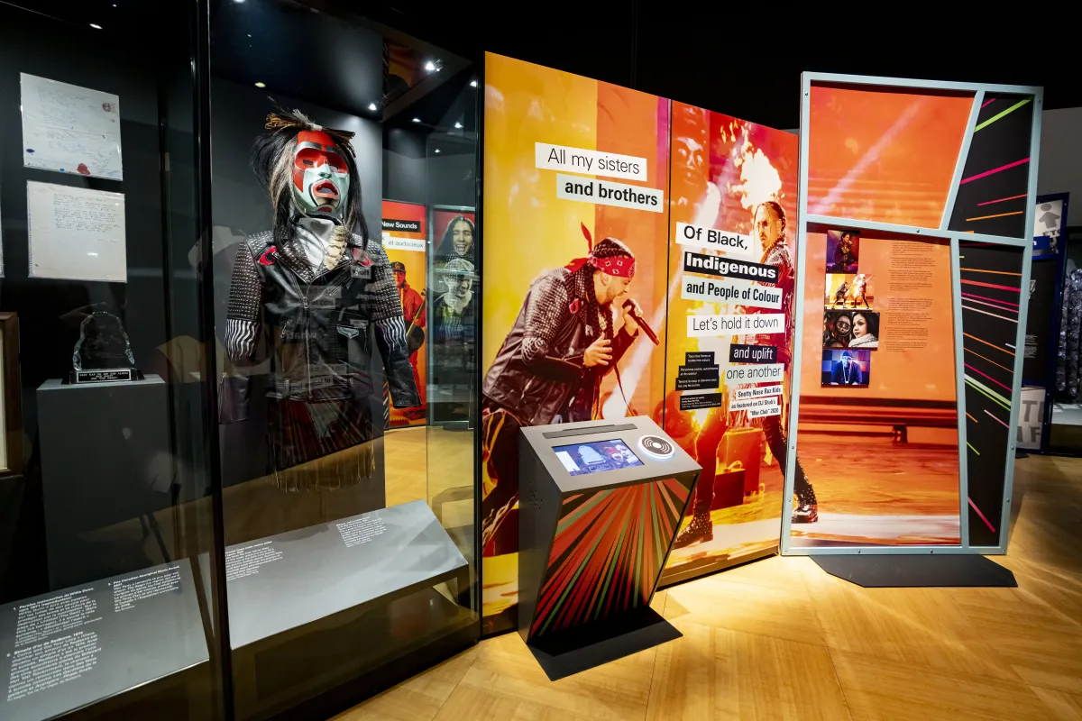 A museum exhibit highlights Indigenous musical artists. It contains a glass case displaying a mannequin wearing a mask and a jacket, as well as a screen on a podium-type structure and large graphic panel of text and images on an orange background. 