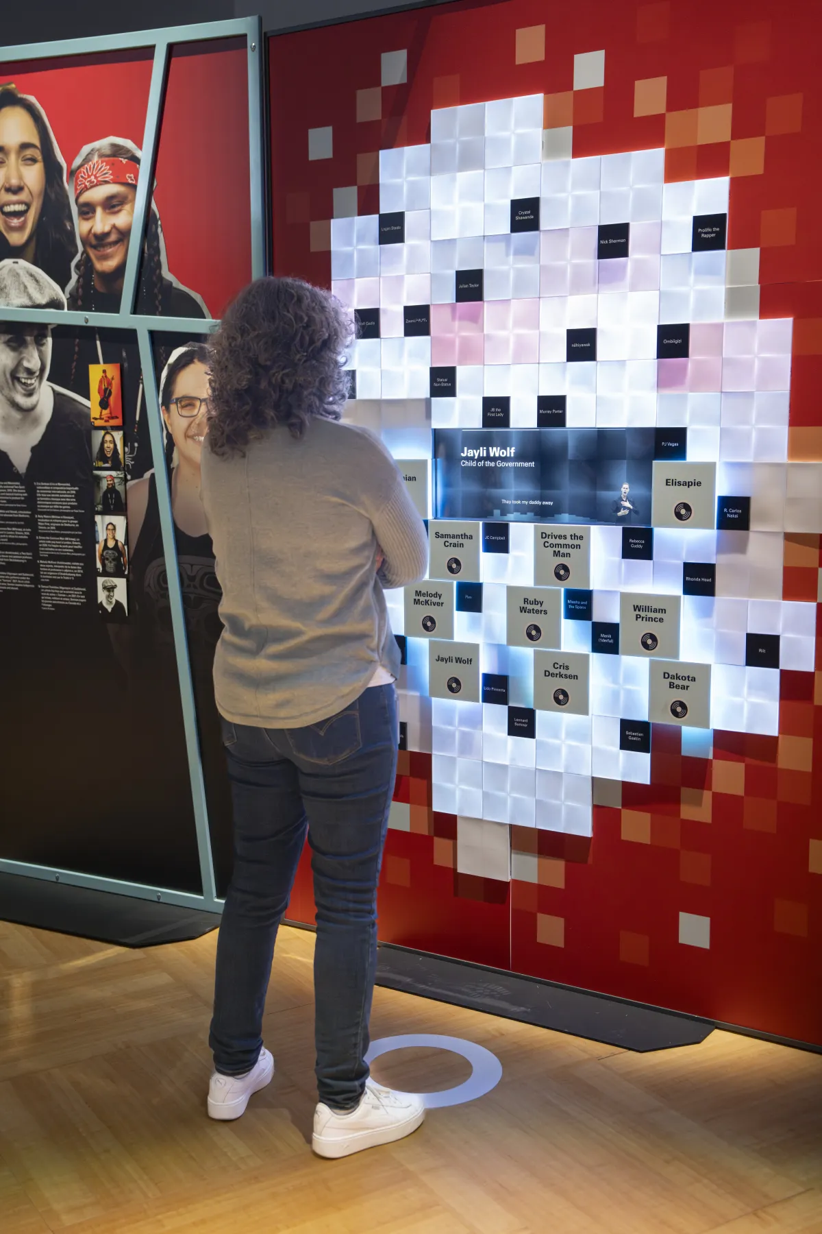 A visitor engages with an interactive wall component made up of small squares with a blue screen in the centre. Light-coloured squares peppered with black ones and grey ones containing text form a circular shape at the centre while squares in shades of red are on the periphery. To the left of the visitor is a graphic panel showing smiling faces.