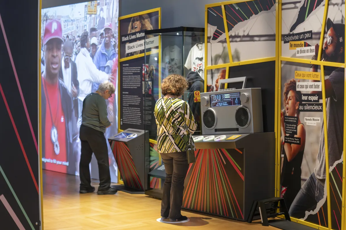 A visitor watches a screen placed on a podium-type structure in a Black Lives Matter exhibit while another visitor interacts with a large radio including a screen displaying text. On the wall in front of them are large graphic panels with images and text.