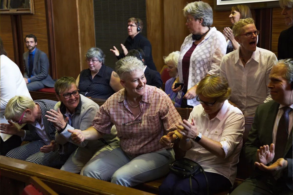 A group of people sitting on benches and standing in a dark wood panelled room. They are clapping and smiling.