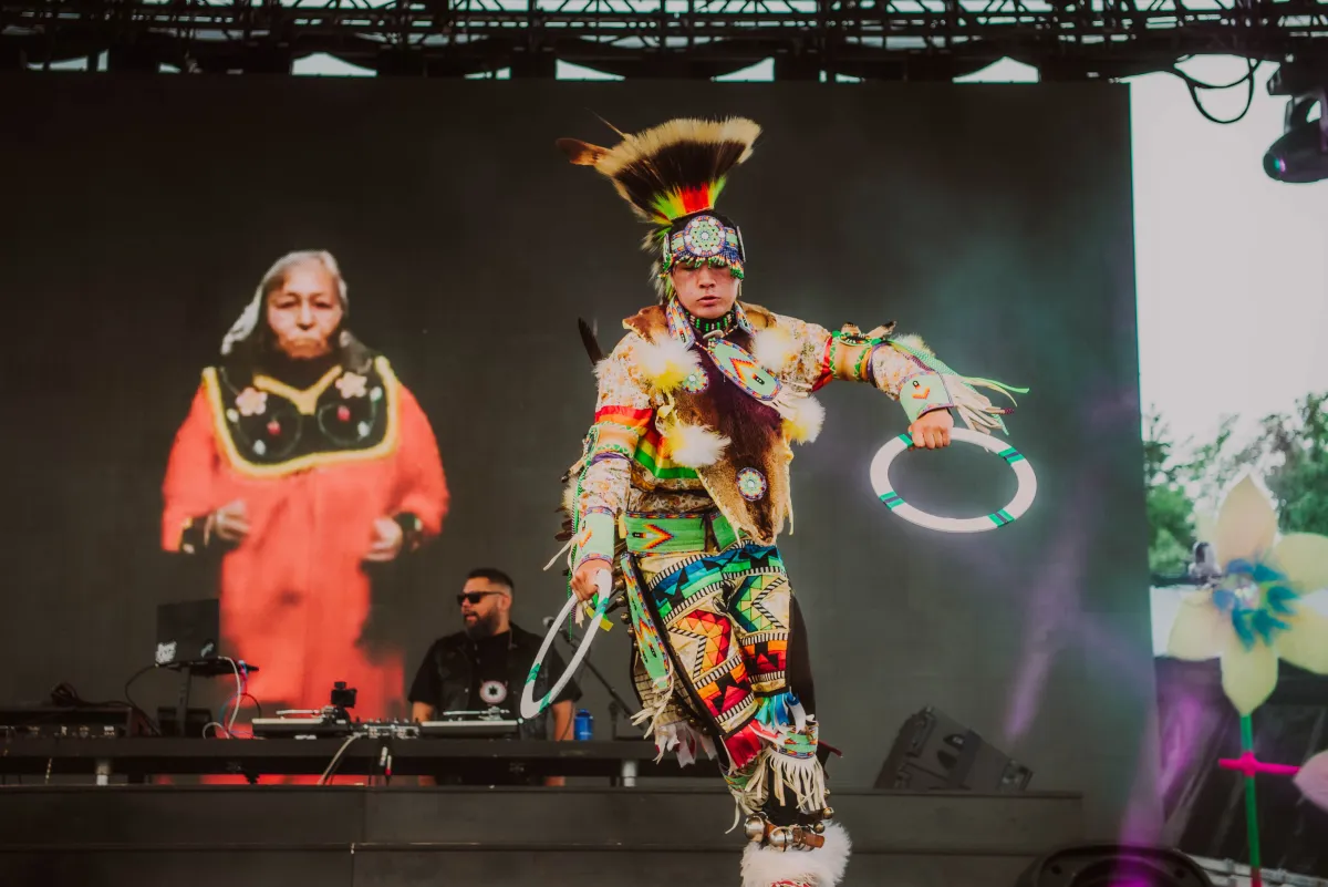 A bearded, dark-haired man wearing black sunglasses spins music on stage with large images of two Indigenous pow wow performers projected on a screen behind him.