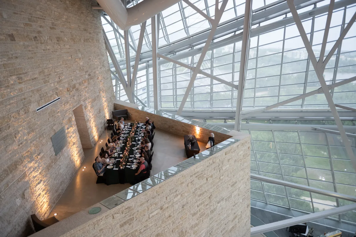 An elegant long table with people dining in an architecturally striking space, featuring tall glass windows and a high ceiling supported by metal beams.