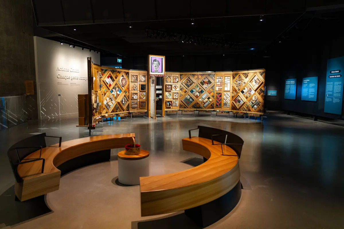 A museum exhibition featuring a large, curved freestanding wooden wall on which is mounted numerous small objects arranged in diamond and rectangular patterns. In the foreground, there circular benches creating an inviting space for visitors to view and contemplate the exhibit.