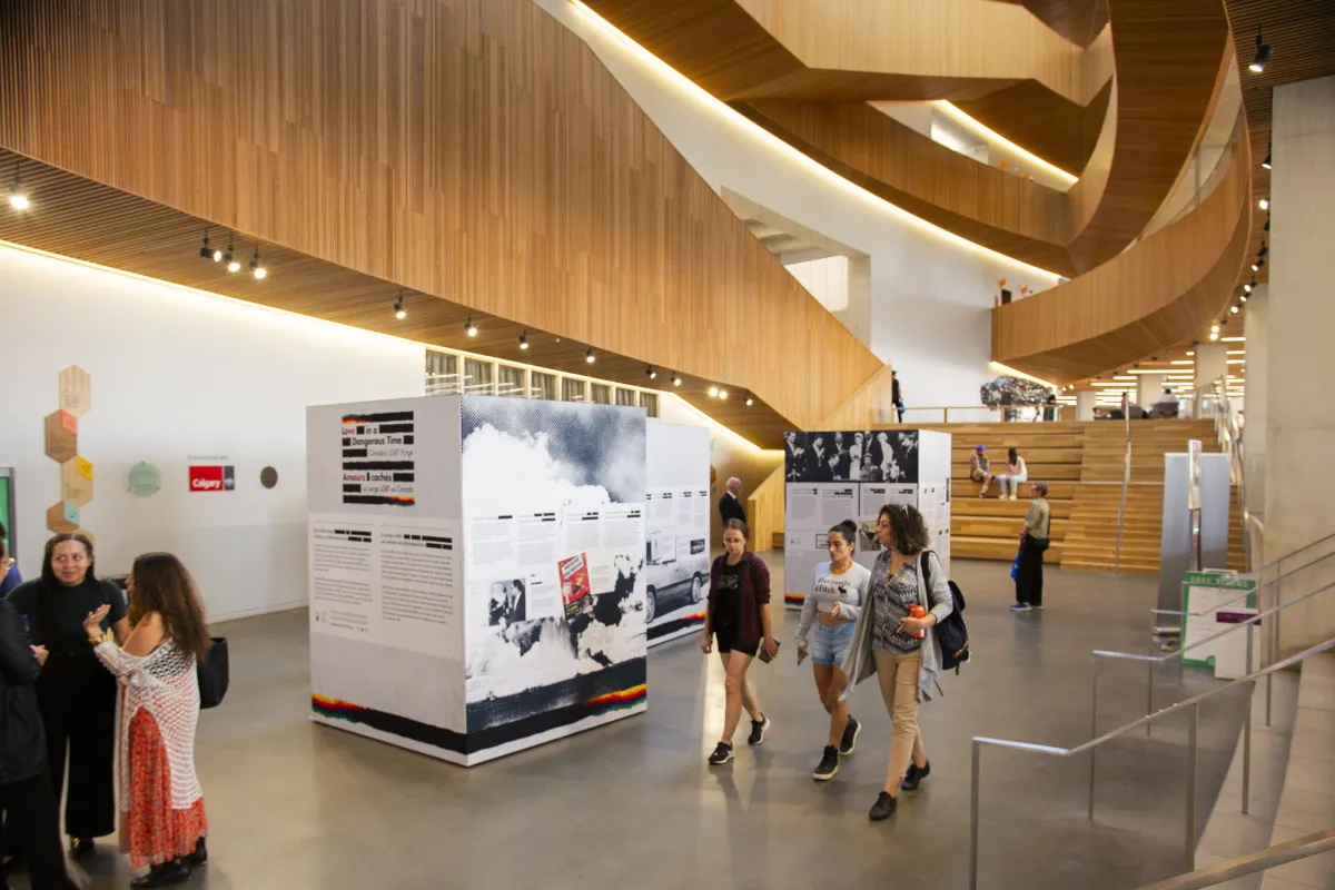 Interior view of a white atrium with wooden levels above. Visitors are walking past three standalone informational displays. The displays include text and images set against a backdrop of wooden architecture and a staircase. Some visitors are engaged in conversation, while others interact with the exhibition.