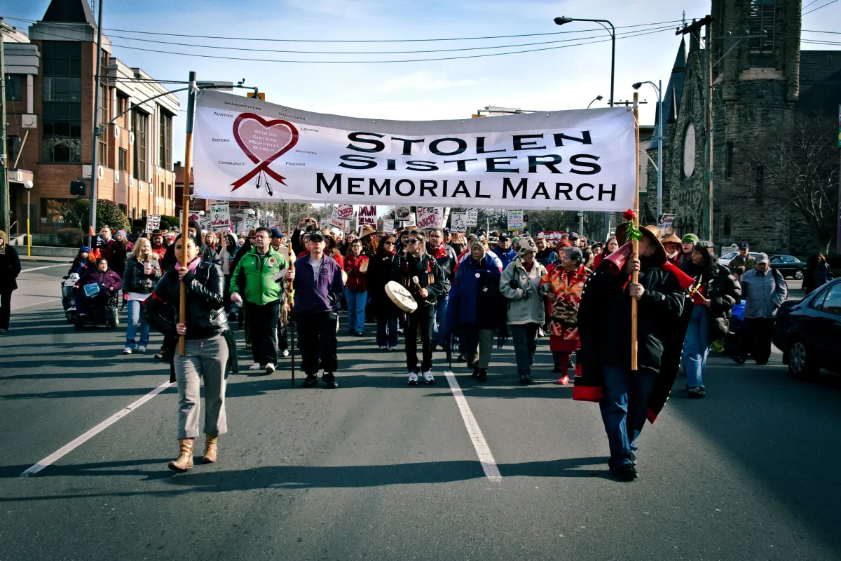 Two women hold a large banner that reads “Stolen Sisters Memorial March” as they lead a large group of demonstrators down a city street.