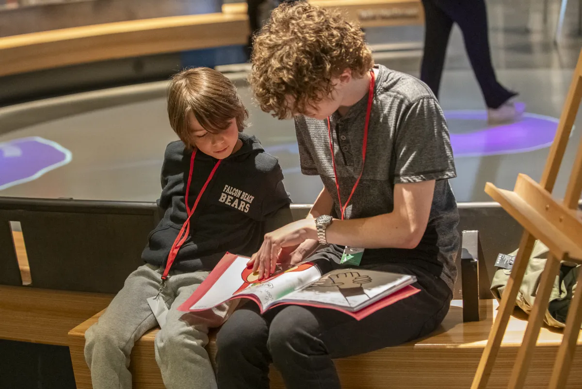 An older and a younger student sitting beside each other on a bench. The older student is reading a book to the younger student. 