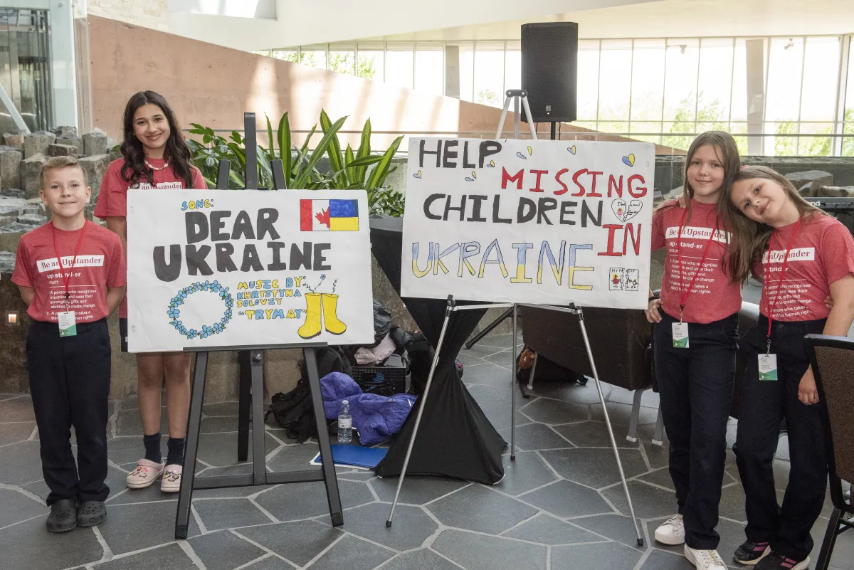 On a sunny day, four students stand on either side of two easels. The students wear matching red shirts that say “Be an Upstander” and the signs on the easels say “Dear Ukraine” and “Help Missing Children in Ukraine.”