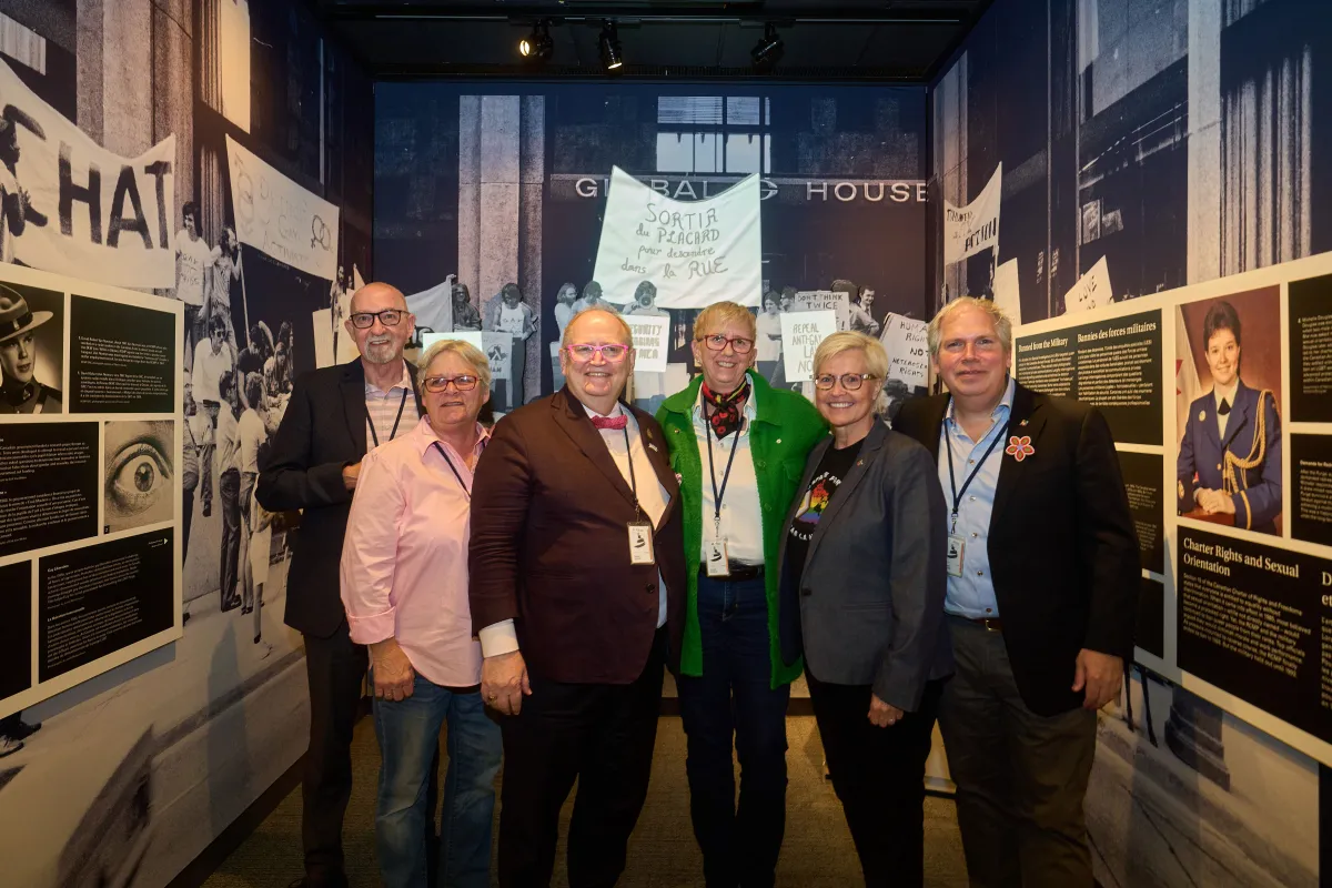 A group of six people face the camera smiling. They are standing in a small space barely large enough to accommodate them. The space is an exhibit made up of words and images.