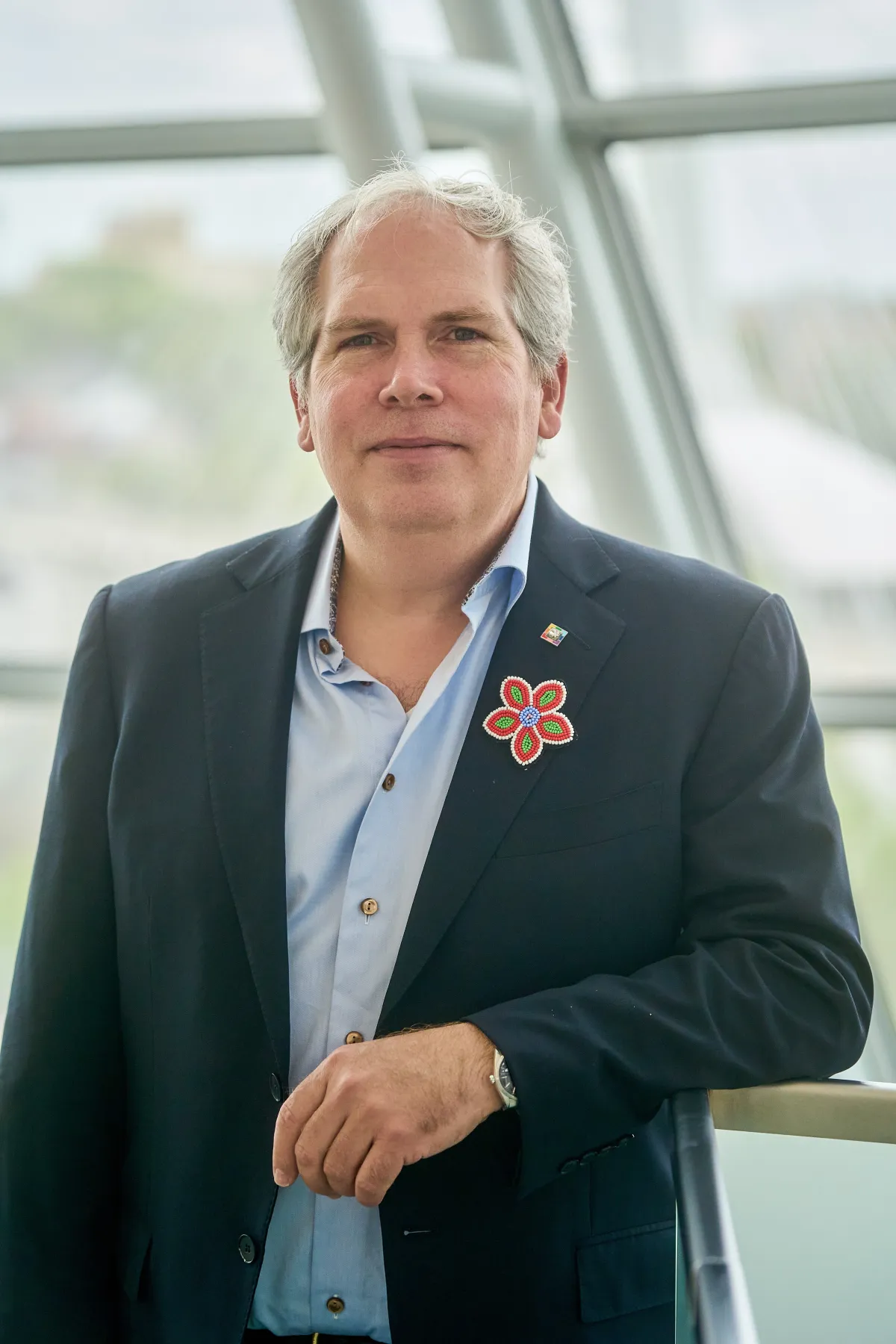 A man stands facing the viewer with a serious expression. He is wearing a dark suit jacket and a light blue buttoned-up shirt. He sports a multicoloured Métis-style flower on his lapel.