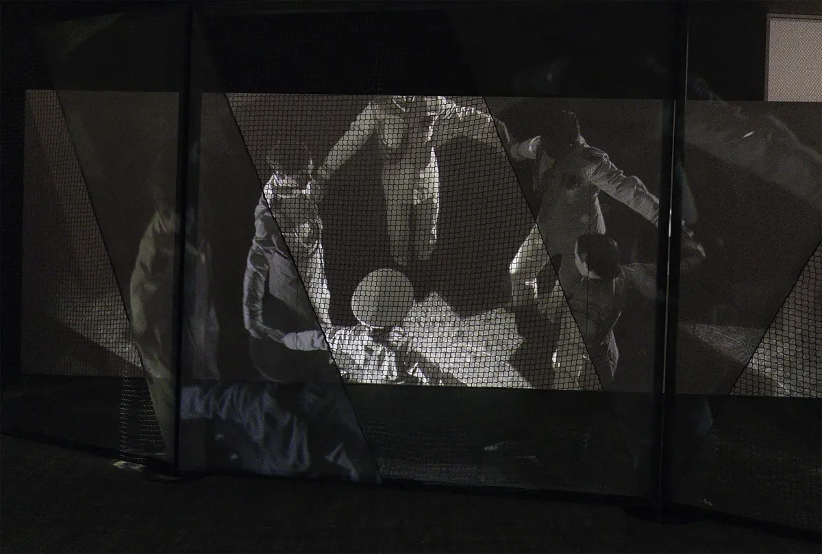 A still from black-and-white film, seen through a heavy black net, depicts dancers dressed in military uniforms forming a circle, holding hands.