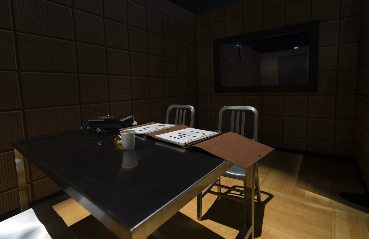 A foreboding, dimly lit space contains two metal chairs and a metal table on which a file and a coffee cup sit.
