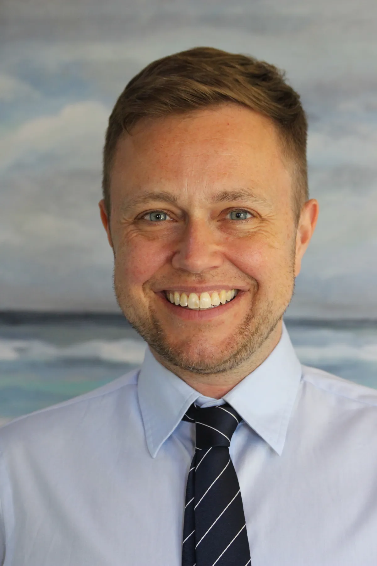 A white man with dark blond hair smiles widely into the camera with waves and a cloudy sky behind him. He is wearing a collared white shirt and a dark tie with diagonal white stripes.