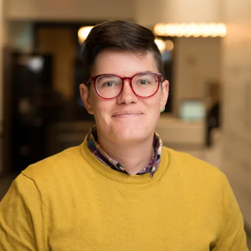 A woman with very short dark hair looks into the camera and smiles with her mouth closed. She wears large round glasses with red frames and a mustard coloured sweater over a collared plaid shirt. The room behind her is blurred.