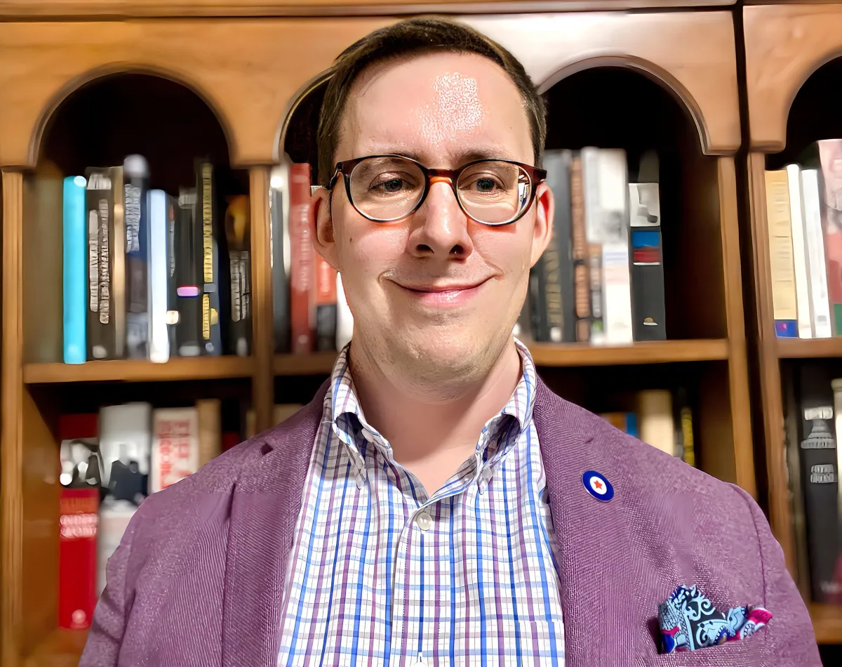 A while man with brown hair and eyeglasses smiles towards the camera. He is wearing a purple blazer with a lapel pin, and a multicoloured dress shirt.