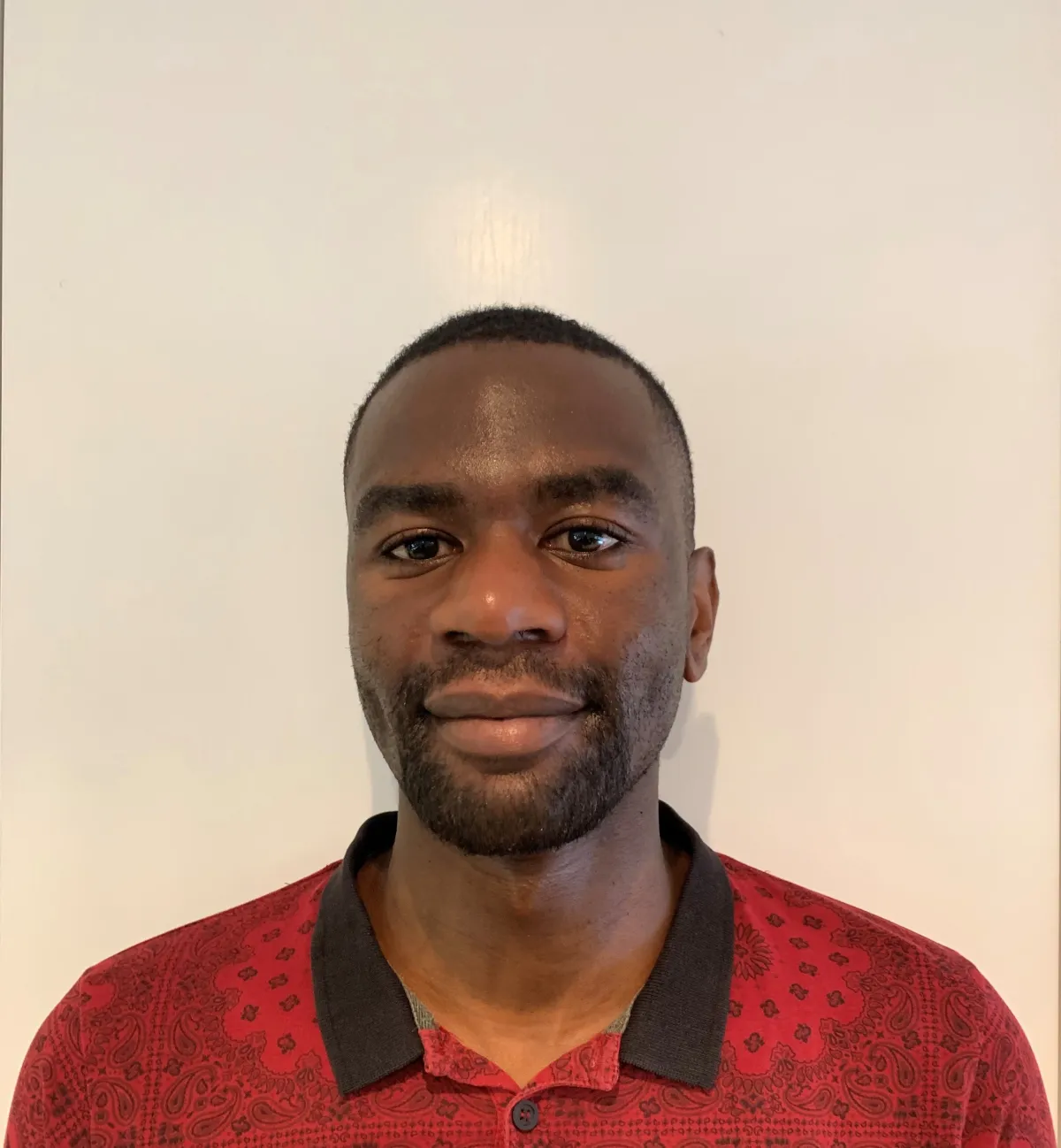 A Black person with very short black hair looks into the camera with a slight smile. They are wearing a red shirt with a printed black line drawing design and a black collar.