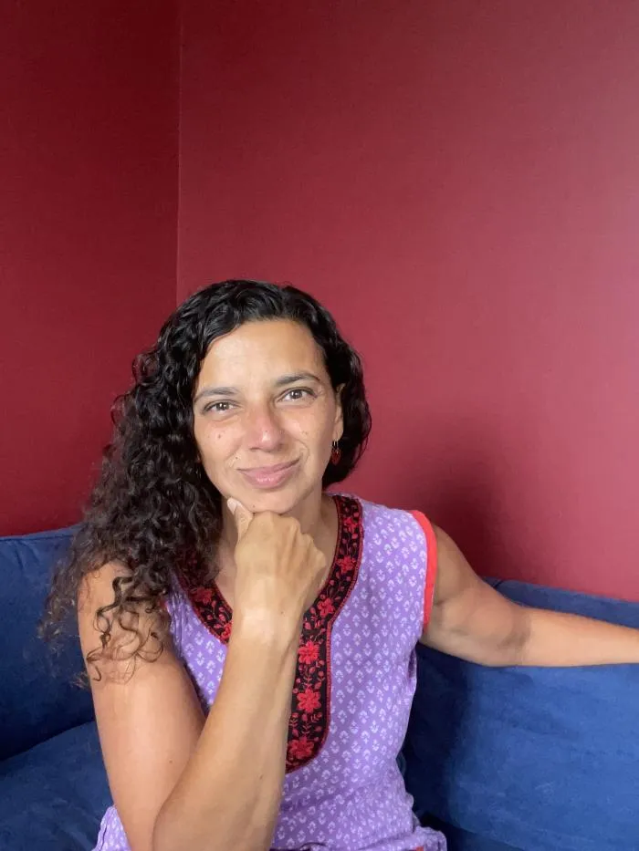 A person with long black curly hair smiles at the camera. Her chin is resting on her right hand and she is sitting on a blue sofa. The wall behind her is dark red.