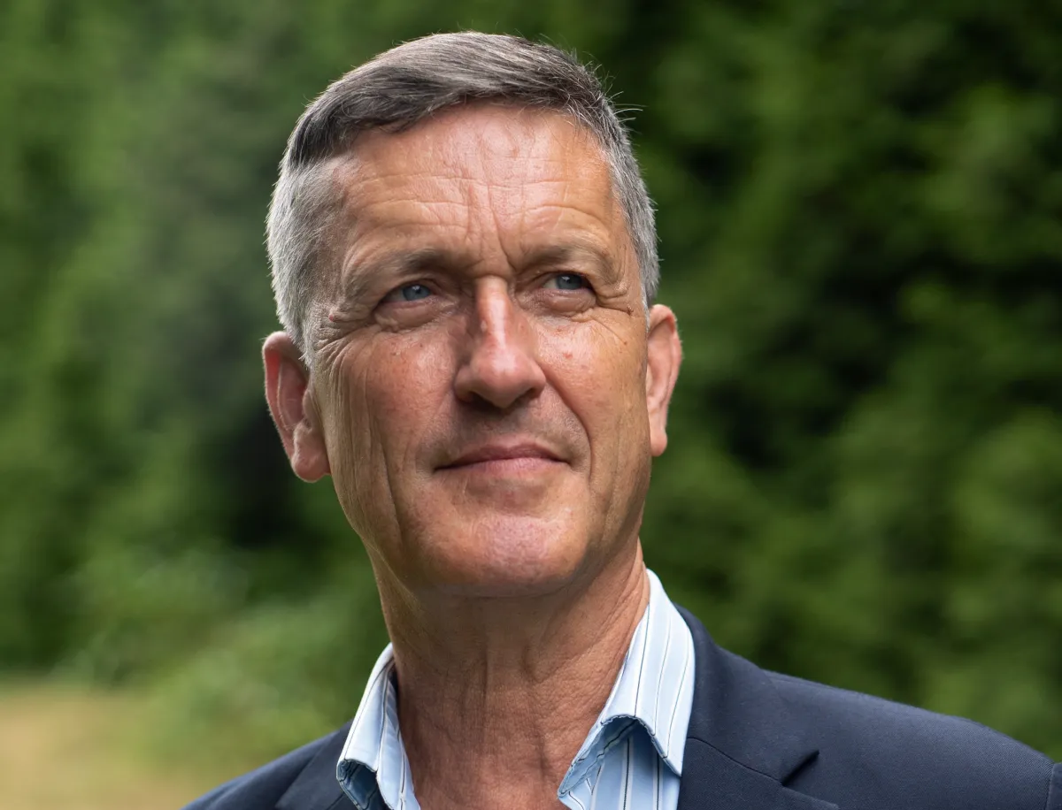 A white male with short grey hair is looking upward away from the camera. He is wearing a blue blazer with a blue striped dress shirt.