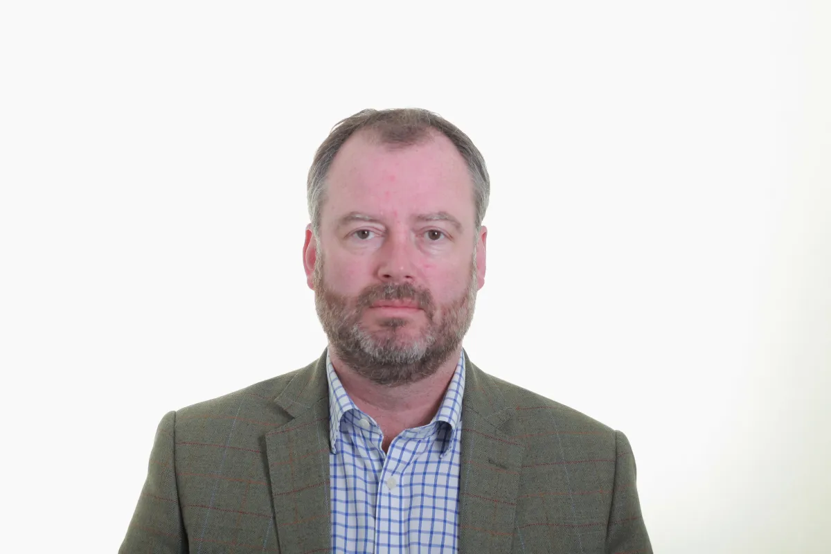 A white man with short greying hair and beard looking impartially at the camera. He is wearing a gray blazer with a blue and white checkered dress shirt.