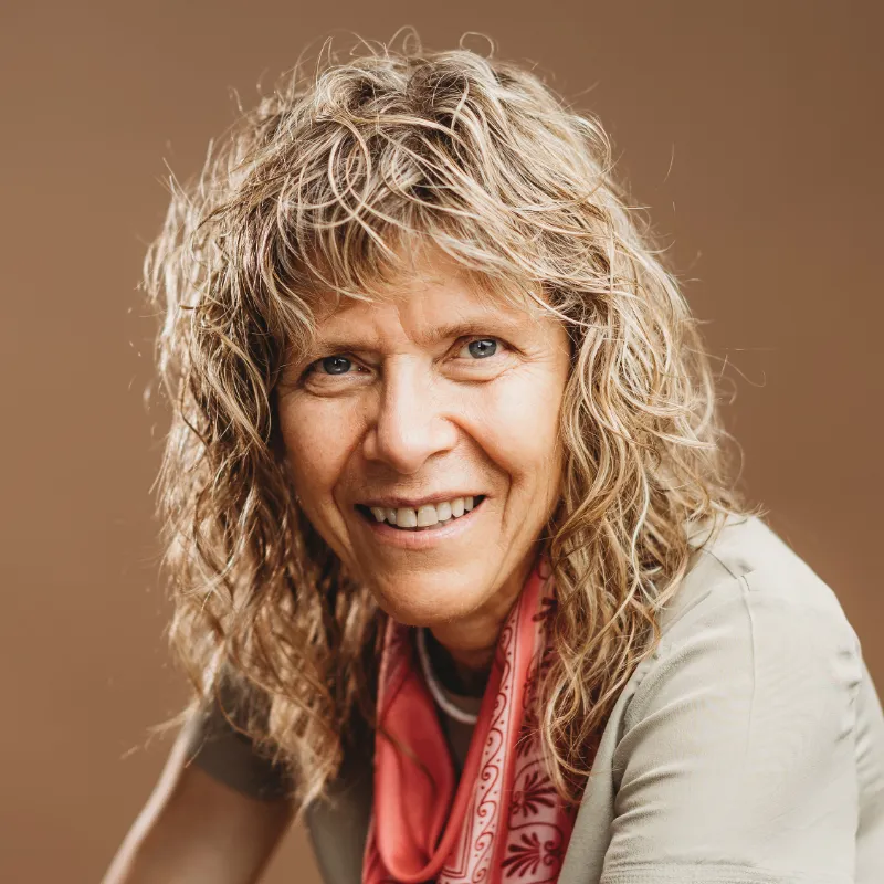 A white-appearing woman with curly blond hair smiles at the camera. She is wearing a beige shirt and a red patterned scarf.