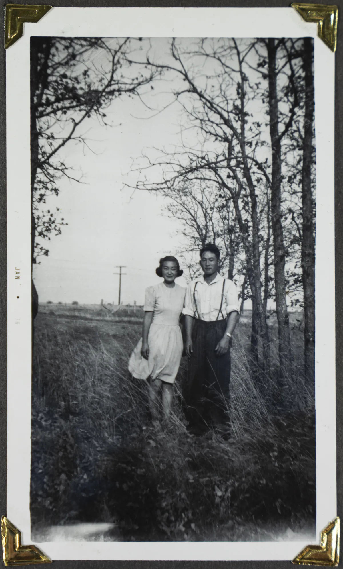  A young woman and a young man pose in a field with trees in the background.