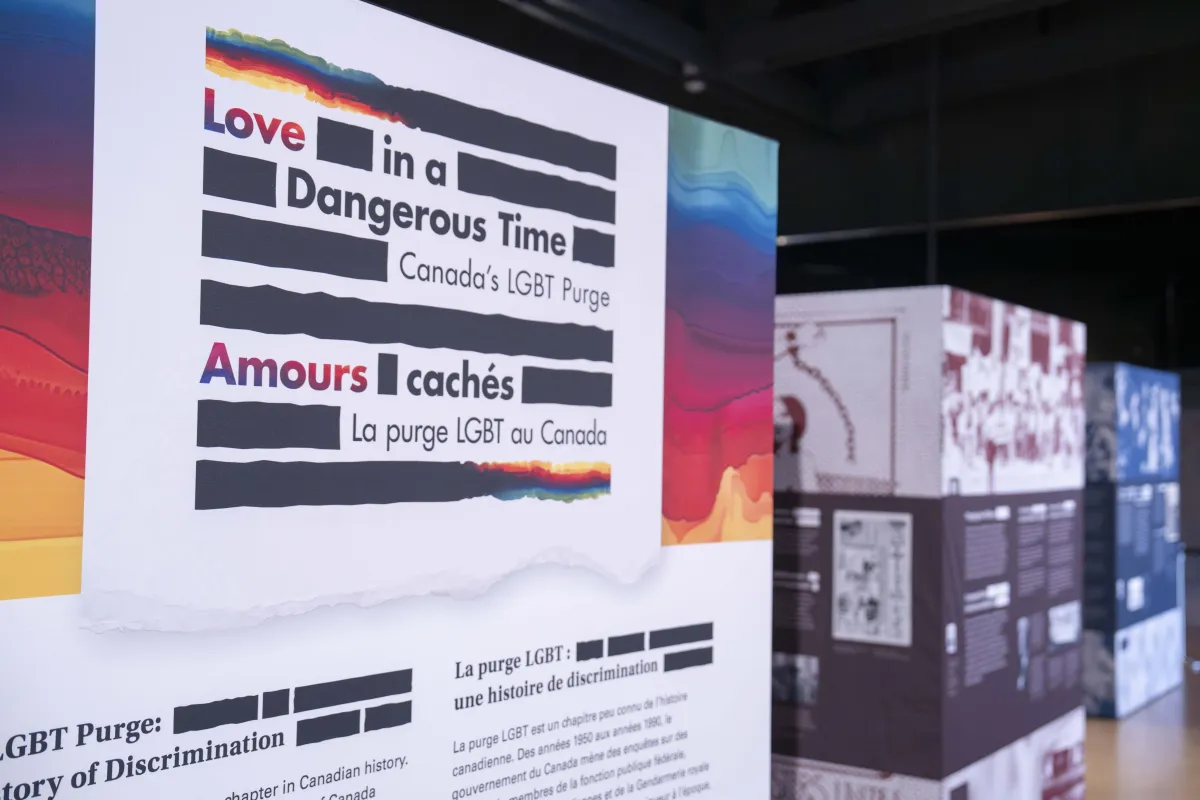 A large sign is made up of thick black horizontal lines and the English and French words “Love in a Dangerous Time: Canada’s LGBT Purge” and “Amours cachés : La purge LGBT au Canada” are distributed within the lines. The top and bottom lines are a mix of black and rainbow colours.
