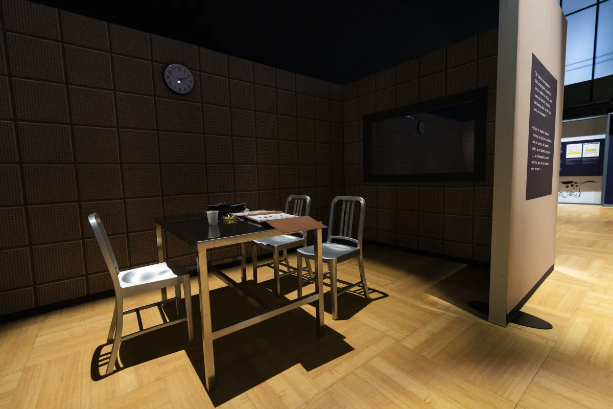 Three metal chairs and a metal table covered with binders and a tape recorder are in the middle of a darkened space. A clock hangs on the wall above the table. A mirrored surface is to the right of the table.