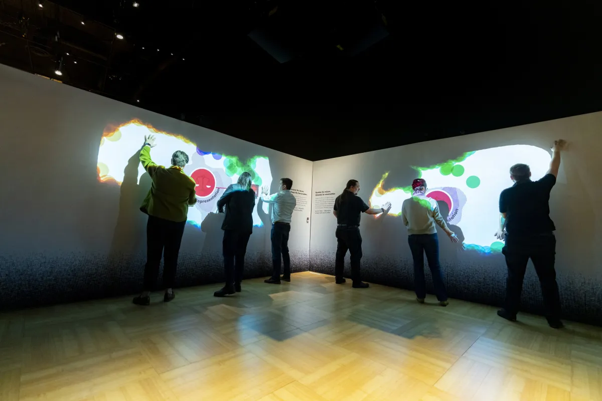 A group of people wave their arms in front of a darkened wall. Colours, shapes, and words appear on the wall where their hands are.