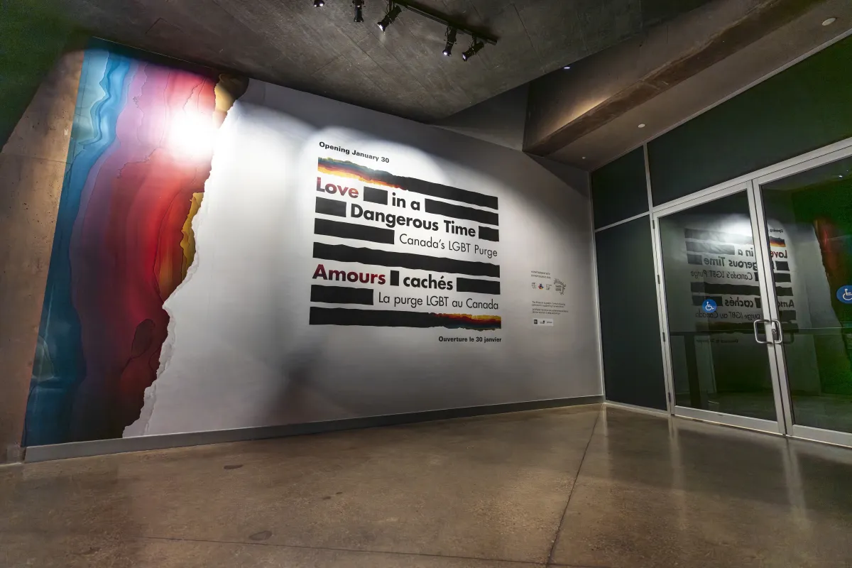 A large sign on a wall is made up of thick black horizontal lines and the English and French words “Love in a Dangerous Time: Canada’s LGBT Purge” and “Amours cachés : La purge LGBT au Canada” are distributed within the lines. The top and bottom lines are a mix of black and rainbow colours. Rainbow colours cover the left edge of the wall.