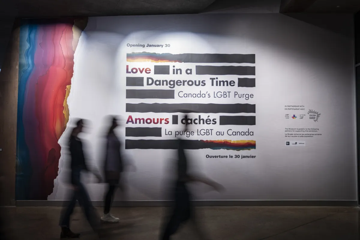 A small group of people walk by a large sign on a wall, made up of thick black horizontal lines with the English and French words “Love in a Dangerous Time: Canada’s LGBT Purge” and “Amours cachés : La purge LGBT au Canada” distributed within the lines. The top and bottom lines are a mix of black and rainbow colours. Rainbow colours cover the left edge of the wall.