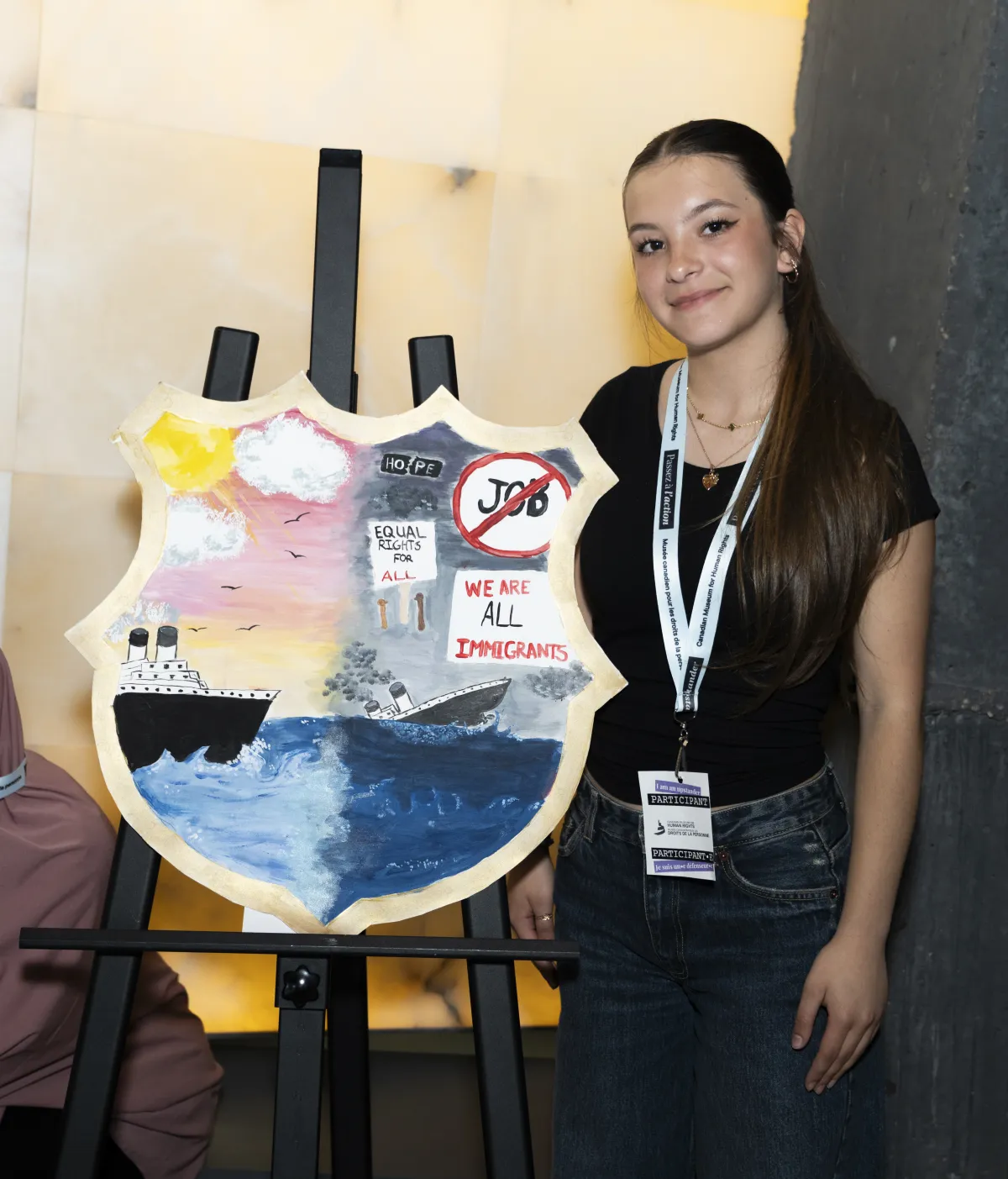 A young woman stands next to a shield painted with sunny skies and a boat floating on water on one side, and a dark sky and a sinking boat.