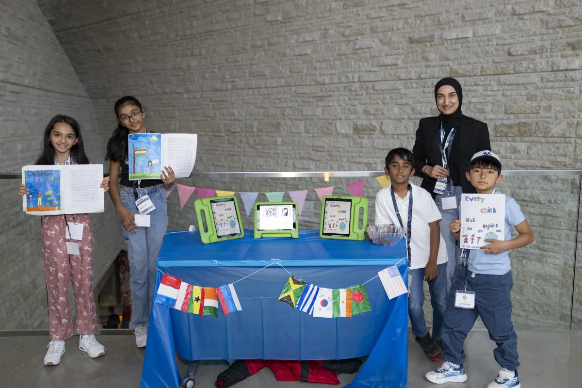 Four students and an adult stand next to a table with flags from countries around the world. Three of them are holding up an open book.