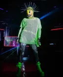 A drag queen wearing a yellow mask with sharp teeth, bright green mesh top and boots, and dark grey tights with large light grey circles.