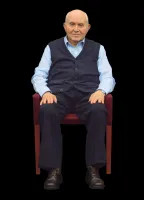 An older man in a light blue shirt and dark blue vest sits on a chair with his hands on his legs. He has a slight smile and is looking directly at the camera.