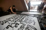 A woman crouches on the floor, painting artistic script on a large white canvas.