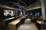 People in a stylish restaurant sitting at tables and long, wooden counters. A metal art installation hangs overhead.