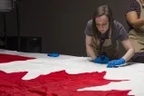 A woman wearing protective gloves and an apron inspects a large Canadian flag, which lies flat on a table.